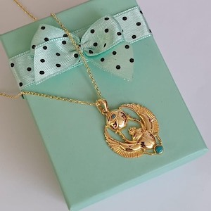 May include: A gold-colored pendant necklace featuring an Egyptian scarab beetle design with turquoise accents. The necklace rests on a light green gift box with a polka-dot bow.