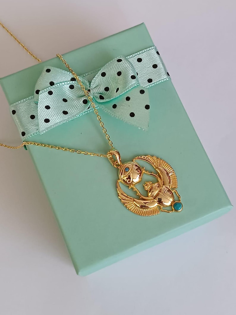 May include: A gold-colored pendant necklace featuring an Egyptian scarab beetle design with turquoise accents. The necklace rests on a light green gift box with a polka-dot bow.