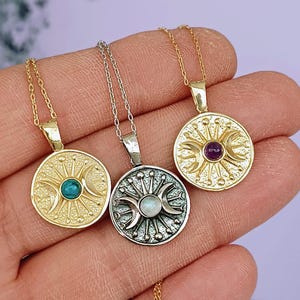 May include: Three celestial-themed pendant necklaces. The pendants are circular with sun and moon designs, and feature gemstones in teal, white, and purple. The necklaces are gold and silver in color, with delicate chain links.