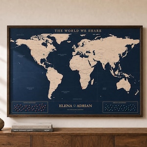 May include: A framed world map print with a dark navy blue background and light beige continents. The text THE WORLD WE SHARE is at the top, and ELENA ADRIAN is centered at the bottom. Two rectangular pin boards are located in the lower corners.