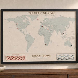 May include: A framed world map titled THE WORLD WE SHARE with a beige background and sage green continents. It features red and blue push pins marking locations, with the names ELENA ADRIAN printed in the center and pin storage areas below.