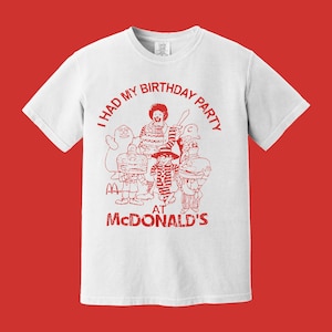 Puede incluir: Camiseta blanca con un gráfico rojo que presenta personajes de McDonald's y el texto "I had my birthday party at McDonald's". El diseño incluye a Ronald McDonald y otras figuras icónicas.