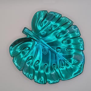 Op de afbeelding: Een teal-kleurige decoratieve bladschotel. De schotel heeft een glanzende, metallic afwerking met een verloop van teal- en blauwtinten. Het bladontwerp heeft gedetailleerde nerven en een textuur.
