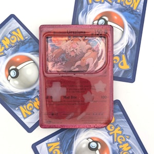 Könnte beinhalten: Eine rote Pokémon-Sammelkarte mit Bloodmoon Ursaluna, mit einem holografischen Bild der Figur. Die Karte ist von anderen Pokémon-Karten mit blauem Hintergrund und einem Pokéball-Design umgeben.