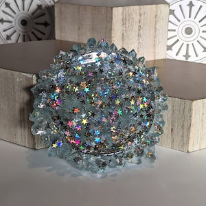 May include: A clear glass coaster with a raised edge and a glittery, iridescent finish. The coaster is filled with small, holographic stars.