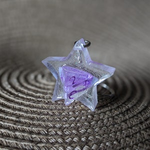 Può includere: Un portachiavi a forma di stella trasparente con un dado iridescente viola e bianco all'interno. Il portachiavi è su una superficie intrecciata marrone.