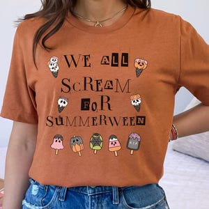 May include: A rust-colored t-shirt with the text "WE ALL SCREAM FOR SUMMERWEEN" in black. The design includes ice cream cones and popsicles. The model is wearing blue jeans.