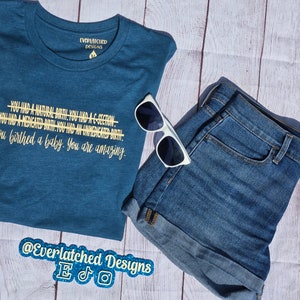 May include: Teal blue t-shirt with the text "You had a natural birth! You had a C-section. You had a medicated birth. You had an unmedicated birth. You birthed a baby. You are amazing." in yellow text. The t-shirt is on a white wood background with a pair of white sunglasses and blue denim shorts.