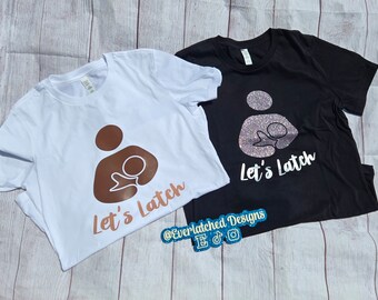 May include: Two t-shirts with the text "Let's Latch" and a graphic of a person breastfeeding a baby. The t-shirt on the left is white with a brown graphic, and the t-shirt on the right is black with a glittery pink graphic. The text "@EverlatchedDesigns" is on the bottom of the black t-shirt.