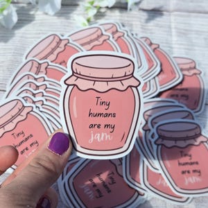 May include: Pink jar-shaped stickers with the text "Tiny humans are my jam." The stickers have a light pink jar with a darker pink lid and a white border. The stickers are on a light wood surface.