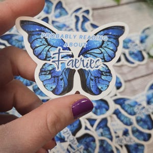 May include: A butterfly-shaped sticker with blue wings and the text "PROBABLY READING ABOUT Faeries". The sticker has a white border and is held by a hand. Other similar stickers are in the background.
