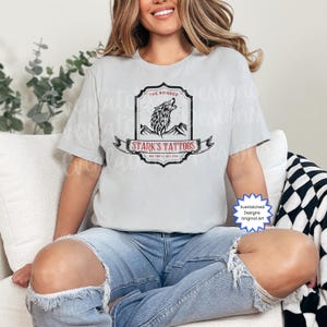 以下が含まれることがあります： 黒と白のグラフィックデザインが施されたライトグレーのTシャツ。デザインには、遠吠えするオオカミ、山、そして「STARK'S TATTOOS」と「THE BONDED」の文字が含まれています。シャツには「ONE YOU'LL KILL FOR」の文字も入っています。