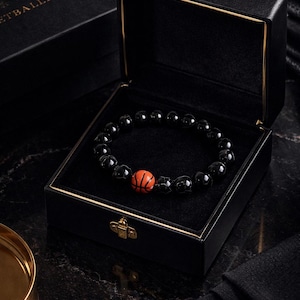 Op de afbeelding: Zwart kralenarmband met een basketbal accent, gepresenteerd in een open zwarte doos met gouden details. De merknaam "ETBALLPLANT" is zichtbaar op de doos. De armband ligt op een zwarte ondergrond.