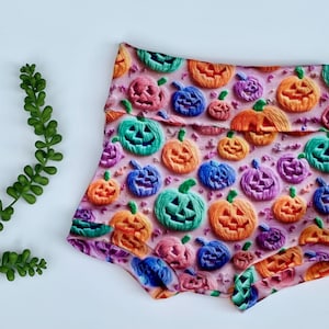 May include: Pink and white patterned baby bloomers with a Halloween theme. The bloomers are covered in colorful jack-o'-lantern faces.