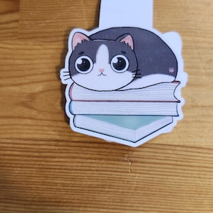 May include: A cat-themed bookmark featuring a cartoon cat with large eyes, resting on a stack of books. The cat is primarily gray and white, with a pink nose. The books are blue and pink.