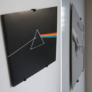 Op de afbeelding: Twee vinylplaten worden met zwarte clips aan een witte muur bevestigd. De plaat links is een zwart album met een regenboog en een driehoek. De plaat rechts is een zwart-wit album met een uitgestrekte hand.