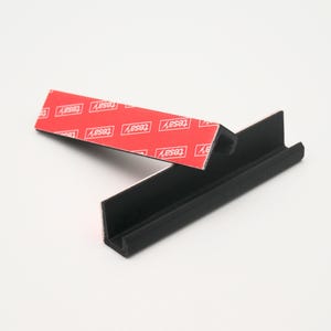 May include: A black plastic corner shelf with adhesive tape on the back. The tape is labeled "TESA" and is designed for mounting the shelf to a wall.