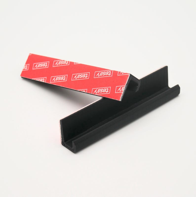 May include: A black plastic corner shelf with adhesive tape on the back. The tape is labeled "TESA" and is designed for mounting the shelf to a wall.