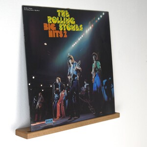 May include: A vintage vinyl record album cover for "The Rolling Stones Hits 2" displayed on a wooden shelf. The album cover features a live concert photo of the band with the album title in orange and yellow text. The record is mounted on a light brown wooden shelf.