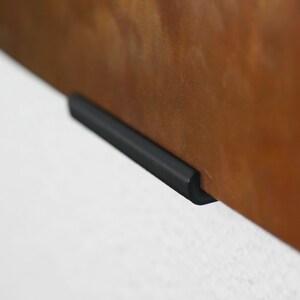 May include: A black metal shelf bracket with a curved edge. The bracket is attached to a rusty metal surface.