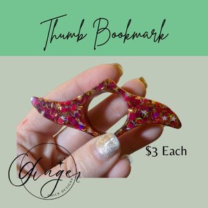 May include: A red and gold glitter thumb bookmark with star-shaped accents. The bookmark has a hole for the thumb and is designed to hold pages open. The text "Thumb Bookmark" is written in cursive.