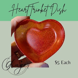 Puede incluir: Un joyero en forma de corazón con un acabado rojo y naranja brillante. El plato se sostiene en una mano y presenta una forma de corazón más pequeña en el centro. El texto "Heart Trinket Dish" está en la parte superior.