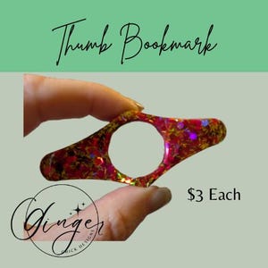 May include: A red and gold glitter thumb bookmark with a large circular opening. The bookmark is held between fingers against a light green and gray background. The text "Thumb Bookmark" is written in cursive.