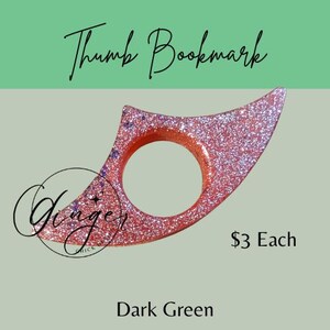 May include: A glittery thumb bookmark in a reddish-pink hue with a circular opening. The bookmark has a pointed, wing-like shape. The text "Thumb Bookmark" is written in cursive at the top. The background is dark green.