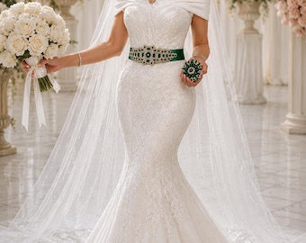 May include: A white lace wedding dress with an off-the-shoulder neckline and a mermaid silhouette. The bride wears a green and gold embellished belt and holds a bouquet of white roses. A sheer veil and tiara complete the look.