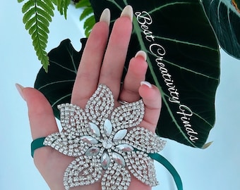 May include: A silver rhinestone flower brooch with a green ribbon attached. The brooch is in the shape of a flower with five petals and a center made of smaller rhinestones.
