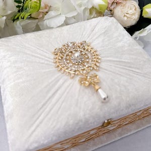 May include: A white velvet ring box with a gold trim and a decorative jeweled ornament on the lid.