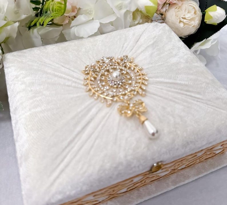 May include: A white velvet ring box with a gold trim and a decorative jeweled ornament on the lid.