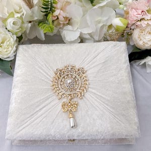 May include: A white velvet box with a gold and crystal embellishment. The embellishment features a round design with a pearl in the centre and a dangling pearl drop.