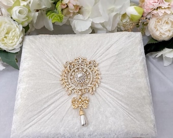 May include: A white velvet box with a gold and crystal embellishment. The embellishment features a round design with a pearl in the center and a dangling pearl drop.