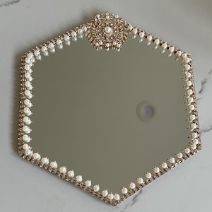 May include: A hexagonal mirror with a gold-colored frame, adorned with faux pearls and rhinestones. The top of the mirror features a decorative embellishment with a pearl center. The mirror's edge is lined with small pearls.