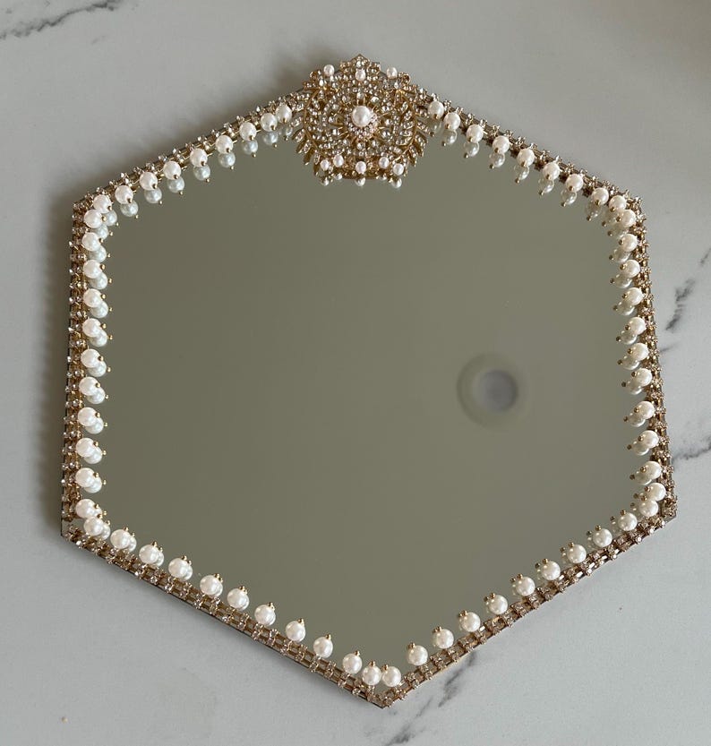 May include: A hexagonal mirror with a gold-colored frame, adorned with faux pearls and rhinestones. The top of the mirror features a decorative embellishment with a pearl center. The mirror's edge is lined with small pearls.