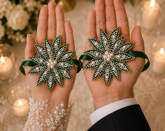 May include: Two beaded floral wrist corsages. Each corsage features a dark green, star-shaped design with white and gold beads, and a central cluster of pearls. A dark green ribbon secures each corsage. The background includes candles and flowers.