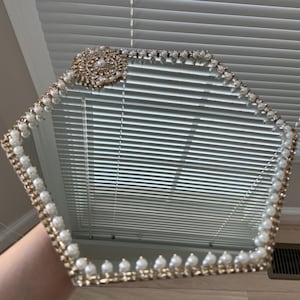 May include: A hexagonal mirrored tray with a gold-colored frame. The frame is embellished with small white pearls and clear crystals. A large, ornate gold-colored floral design is in one corner.