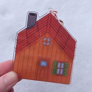 Op de afbeelding: Een transparante acryl sleutelhanger in de vorm van een klein oranje huis met een rood dak, een schoorsteen, een klein raam en een deur.