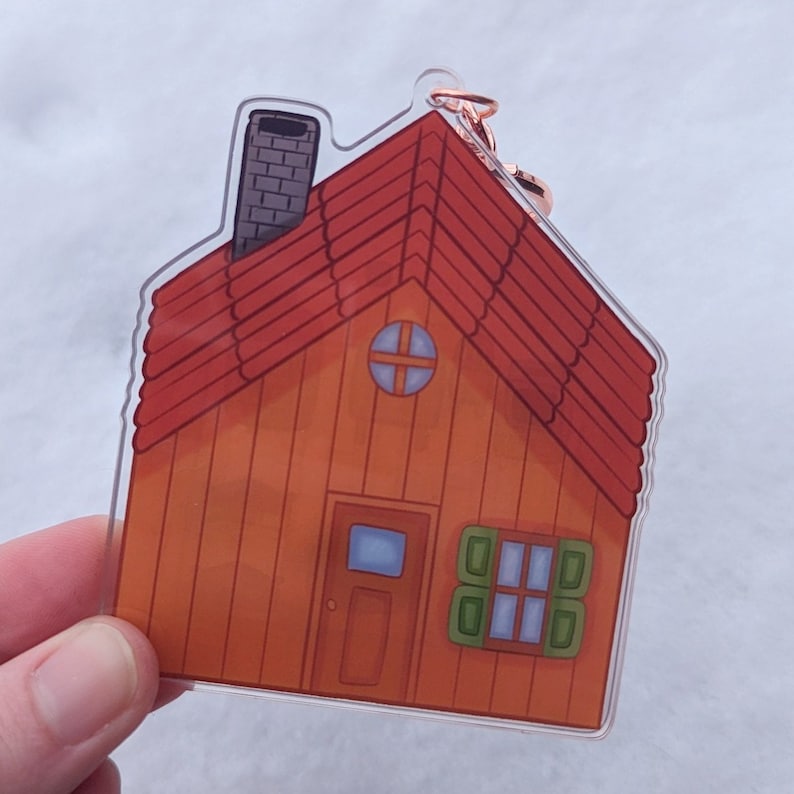 Op de afbeelding: Een transparante acryl sleutelhanger in de vorm van een klein oranje huis met een rood dak, een schoorsteen, een klein raam en een deur.
