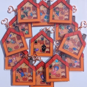 Op de afbeelding: Een set van 10 kleurrijke acryl sleutelhangers met cartoonfiguren in verschillende poses in kleine huisvormige frames. Elke sleutelhanger heeft een gouden metalen ring en ketting.