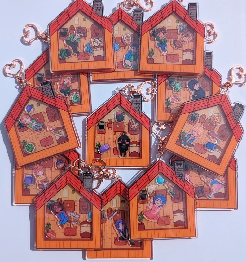 Op de afbeelding: Een set van 10 kleurrijke acryl sleutelhangers met cartoonfiguren in verschillende poses in kleine huisvormige frames. Elke sleutelhanger heeft een gouden metalen ring en ketting.