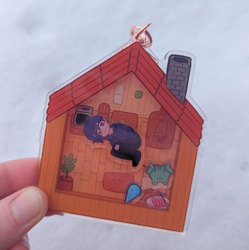 Op de afbeelding: Een transparante acryl sleutelhanger in de vorm van een huis met een rood dak. Binnen in het huis is een cartoonfiguur met blauw haar en een blauw shirt. Het figuur zit op de vloer van het huis. Er zijn andere cartoondetails in het huis, waaronder een plant, een open haard en een tafel met eten.