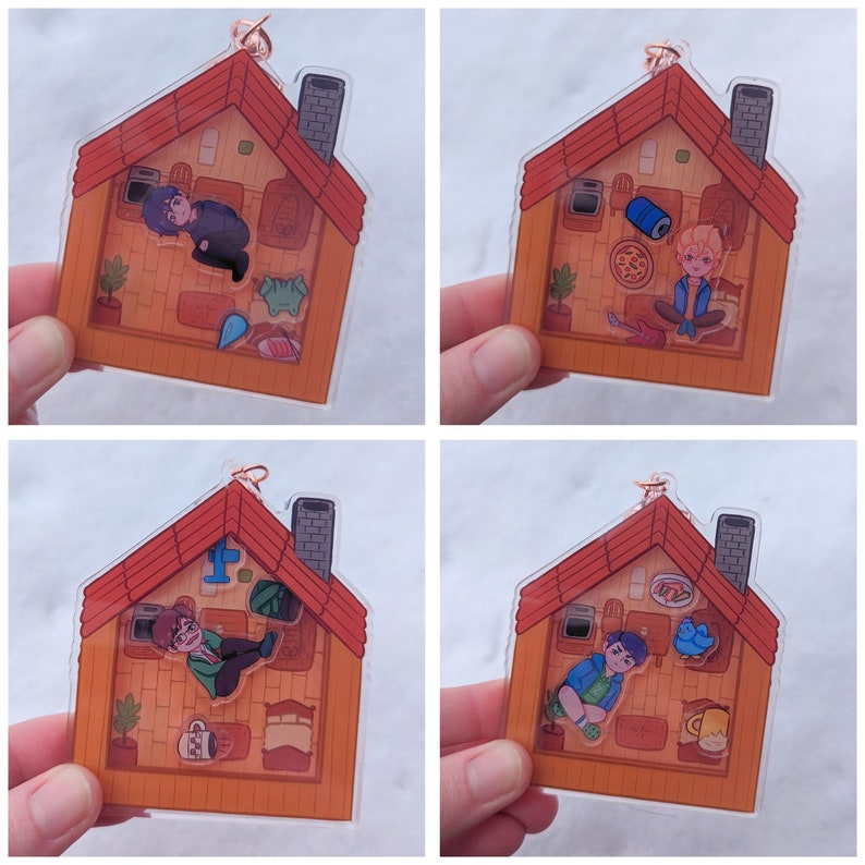 Op de afbeelding: Vier verschillende ontwerpen van transparante acrylcharms met een cartoonhuis met een rood dak en verschillende personages erin. De charms zijn ongeveer 5 cm groot.