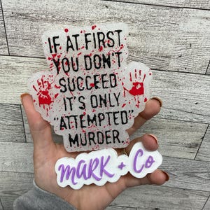 May include: White, glittery, resin coaster with red blood splatter and black text that reads "If at first you don't succeed it's only "attempted" murder."  A smaller white, glittery, resin coaster with purple text that reads "Mark + Co" is in the foreground.