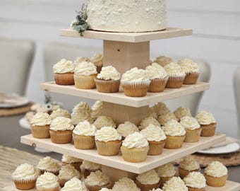 May include: A four-tier wooden cupcake stand with a white cake on the top tier. The stand is decorated with cupcakes with white frosting.