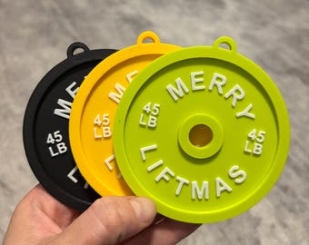 May include: Three colorful weightlifting plate ornaments. The ornaments are black, yellow, and green. Each plate has the text "MERRY LIFTMAS" and "45 LB" in white. Each plate has a small loop at the top for hanging.