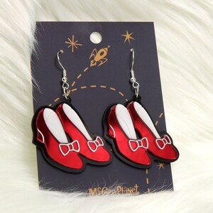 May include: A pair of red ruby slipper earrings with silver hooks. The earrings are made of acrylic and have a shiny finish. The earrings are on a black card with a rocket and stars design. The card also has the text "McGee Planet".