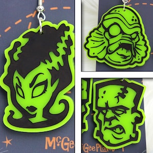 May include: Three green and black Halloween-themed earrings. The earrings feature a monster, a creature from the black lagoon, and a female monster. The earrings are made of acrylic and have a black outline.