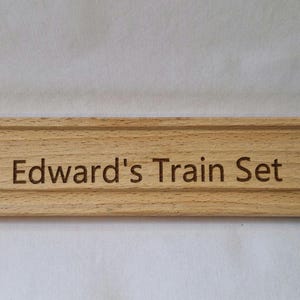 May include: A light brown wooden train track piece, engraved with the text "Edward's Train Set". The track has a grooved design for train wheels and features a rounded end and a notch for connection. The track is approximately 20 cm long.